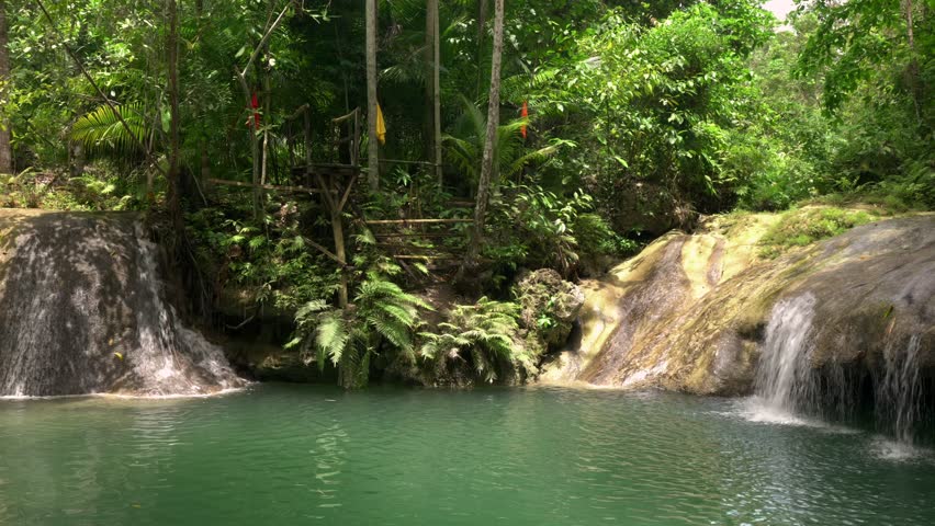 Stationary camera, man swims across the pool, climbs a waterfall with a rope, then does a somersault into the water from a jumping platform.