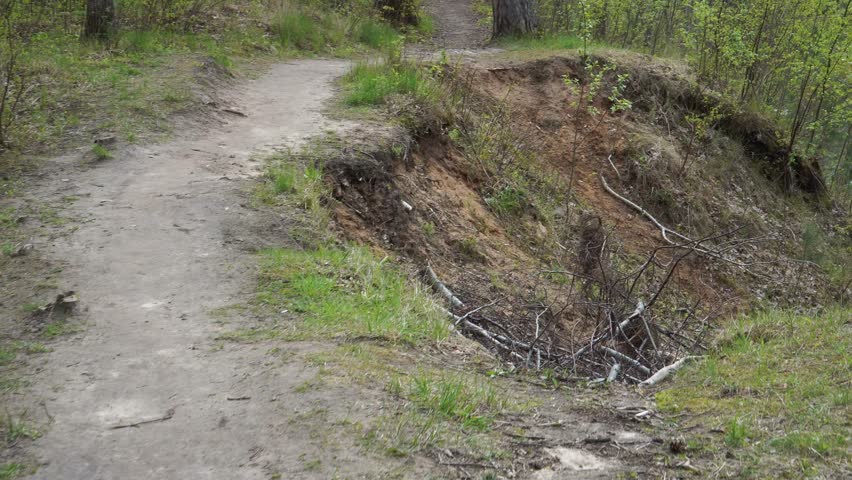 Camera moves dynamically toward an eroded sandy cliff above a spring river. The view tilts down a steep slope revealing fallen trees, scattered branches, and a partially blocked stream below.