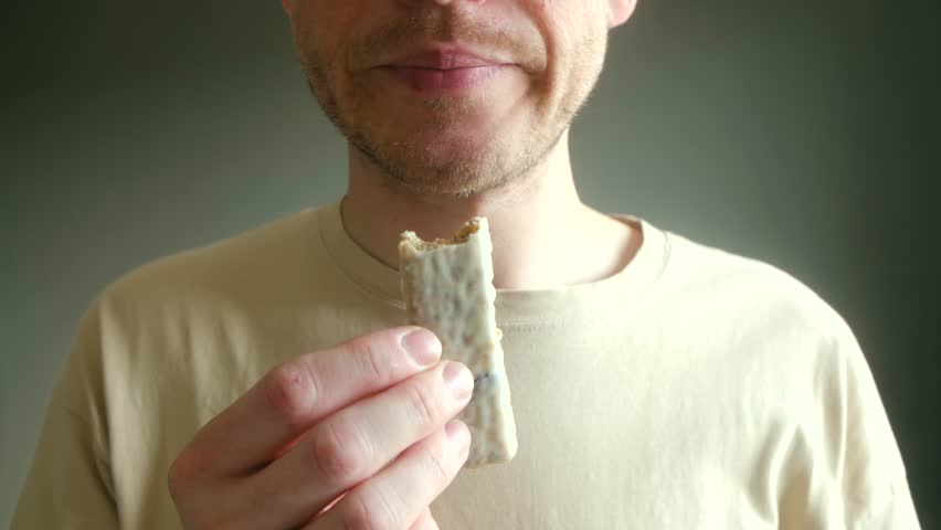 Unrecognizable caucasian man eating white chocolate protein bar for quick snack, enjoying healthy and nutritious meal replacement for energy and muscle recovery. Unrecognizable man eating protein bar