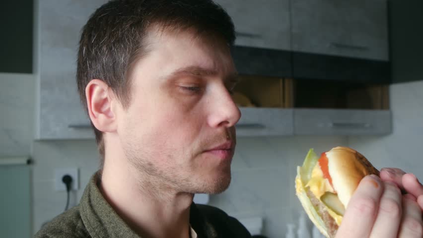 Hungry young caucasian man greedily taking large bite of fresh, juicy hamburger with cheese and pickles, chewing with great appetite and satisfaction in his kitchen. Hungry man biting big hamburger