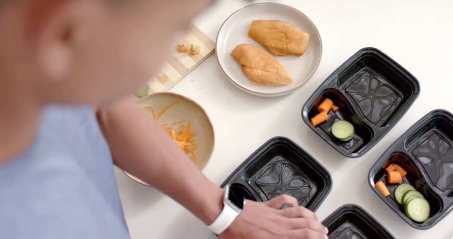 Hand starting meal prep on countertop, slicing cucumber and filling black trays for lunches. Kitchen, containers, organization, overhead, utensils, vegetables, planning