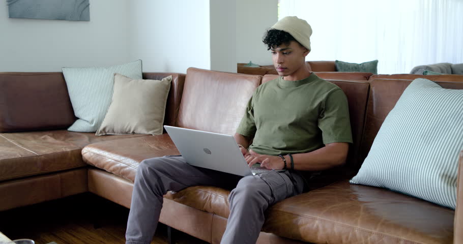 African American man using silver laptop on couch, smiling at message, leaning forward, copy space. Remote, work, productivity, device, typing, focus, daylight