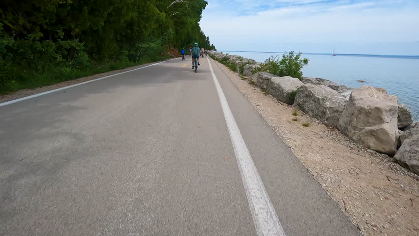 POV Biking on M-185 with Sailboat on Lake Huron, Mackinac Island, Michigan, USA. A slow-motion, point-of-view shot from a bicycle traveling along M-185 on Mackinac Island, Michigan. The footage features a large group of cyclists in the distance on the paved coastal road and a white sailboat cruising on the blue waters of Lake Huron under a Summer sky.