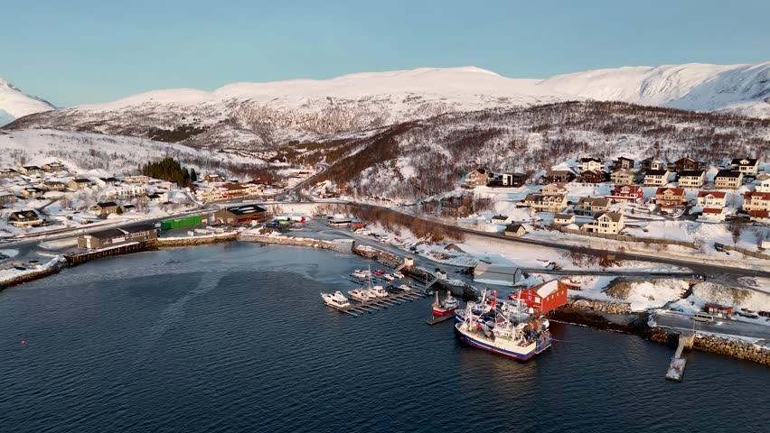 Drone panoramic view over coastal harbor and residential area near Tromsø, Norway. Fishing boats, waterfront buildings and snow covered Arctic mountains in winter daylight.