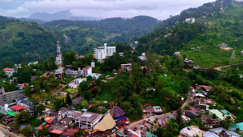 Aerial view of Ella town nestled in green hills with houses and winding roads under a cloudy sky, showcasing lush vegetation and mountain scenery.