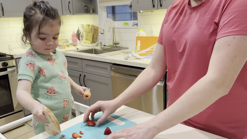 Mother and 1.5-year-old toddler preparing fresh strawberries together in a modern home kitchen using toddler-friendly food preparation tools. Authentic lifestyle moment showing bonding, early learning, and child independence as the toddler practices cutting fruit with gentle parental guidance. Handheld mid-range footage captured in natural light emphasizing Montessori-style learning and real family interaction.
