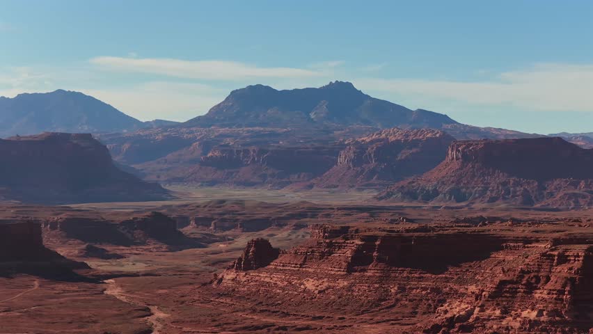 A sweeping drone perspective captures the monumental scale of layered crimson canyons and distant mountain peaks. This Utah desert landscape offers a majestic, location-agnostic backdrop for high-conc