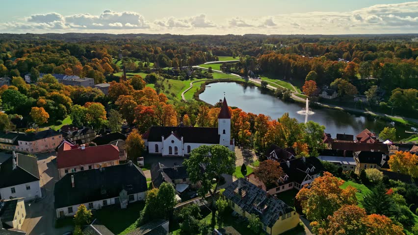 Aerial view of white church with red steeple beside calm lake and fountain, surrounded by autumn village in Europe