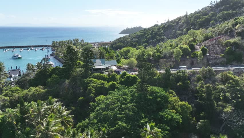 Slow drone pullback and rise in the afternoon, revealing Hayman Island’s marina, resort lagoon pool and turquoise bay framed by lush hillside and sunlit ocean, Whitsundays, Queensland, Australia.