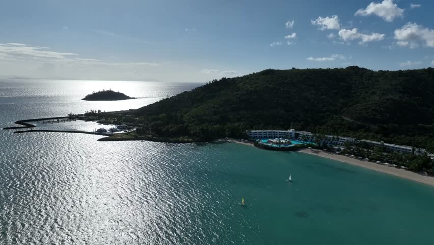 Slow drone pan right in the afternoon, sweeping across Hayman Island’s sunlit bay toward the beachfront resort and lagoon pool with the marina and offshore islet in view, Whitsundays, Queensland, Aust