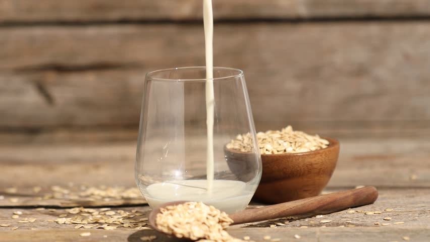 Pouring oat milk into glass and flakes at wooden table, closeup