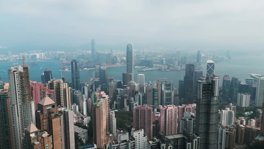 Aerial scenic drone footage of downtown Hong Kong viewed from Victoria Peak on a sunny day, showcasing the skyline, harbor, and dense urban landscape.