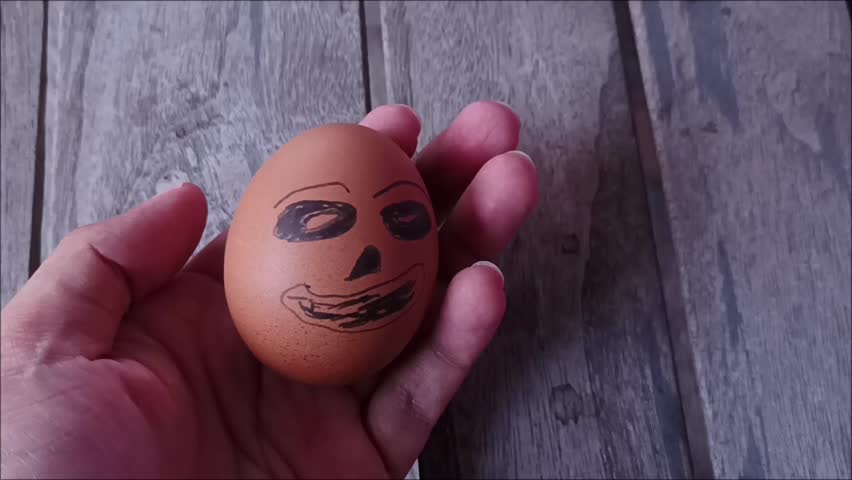 A human hand holds a brown Easter egg decorated with a cute, creatively hand-drawn expression. Wooden background.