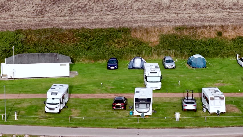 Drone footage shows recreational vehicles and tents parked on a green field near a building.