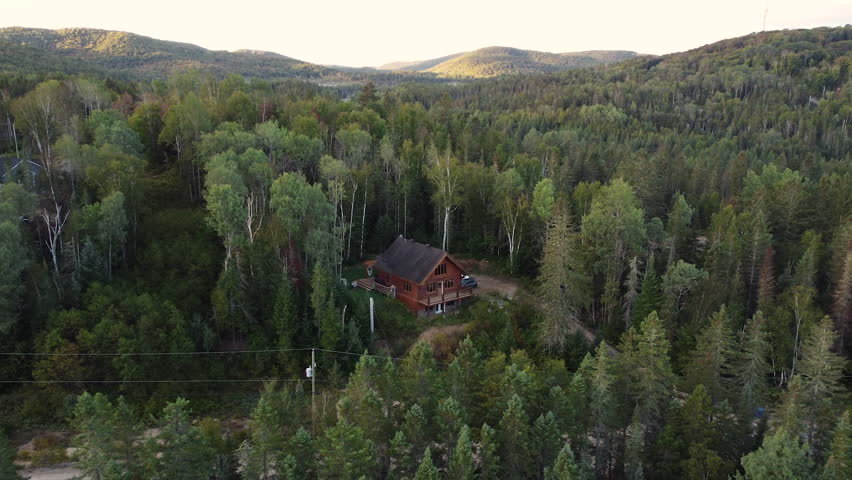Establishing shot of cabin. A drone flies down towards a single wood cabin surrounded by trees, vast forest and mountains in the background. A peaceful serene scene to escape.