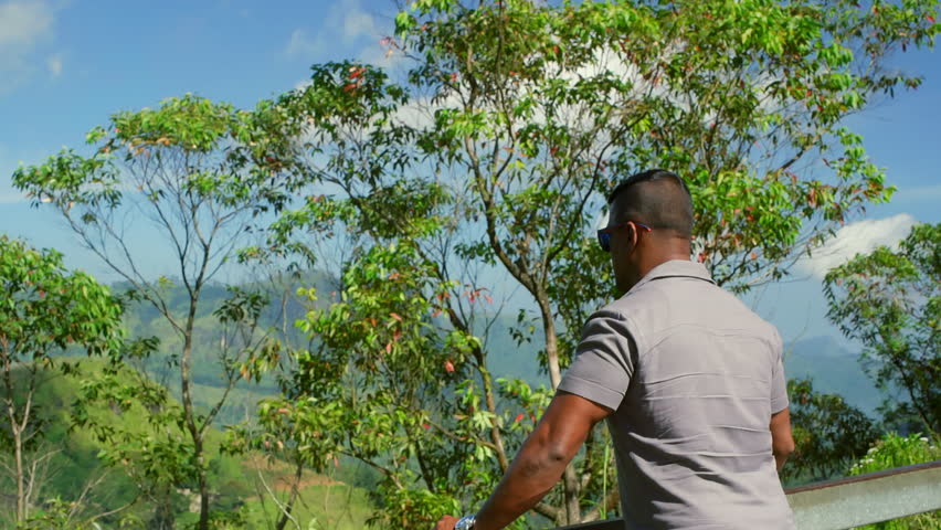 A man stands on a viewpoint, looking out at a stunning, lush green mountain range in a tropical location. A calm, contemplative scene of travel, nature, and inspiration.