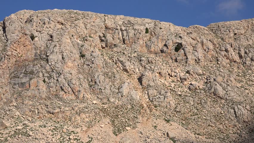 Rocky mountain ridge with tall cliff wall and jagged peaks under bright clear sky. Stony highland escarpment reveals crags, fractured summit chain across barren hillsides, daylight.