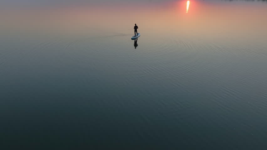 Silhouette of a man fishing on a stand up paddle board at sunset, calm water reflection, outdoor workout and modern healthy lifestyle concept with peaceful nature background.