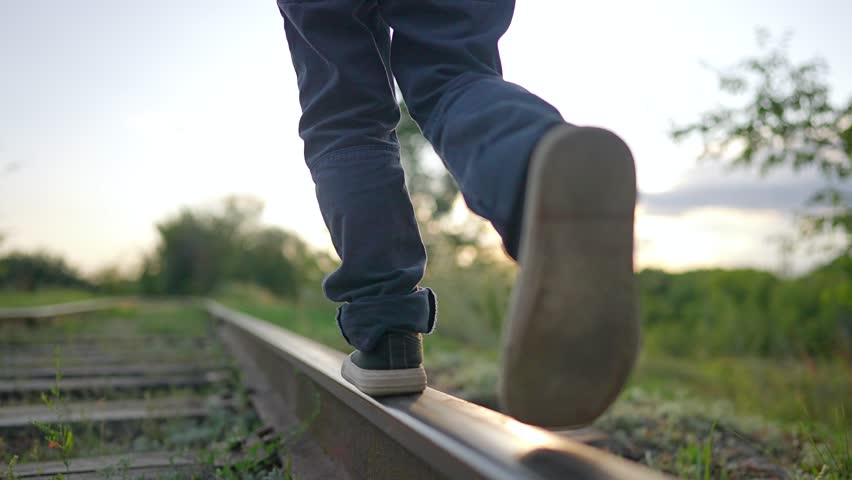 Close up of feet walking on metal railroad track. Child in sneakers balances on railway rail. Legs walk along train track in summer sunset. Kid walking on rail enjoys fun nature journey outdoors.
