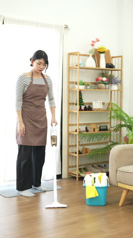 Asian lady doing house chores in apron. young housewife using vacuum cleaner cleaning the wooden floor in the living room.