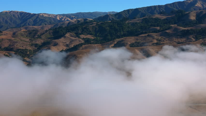 Cinematic drone perspective over rugged Big Sur California coastline where rolling hills emerge from thick coastal fog along the Pacific Ocean under vibrant blue sky.