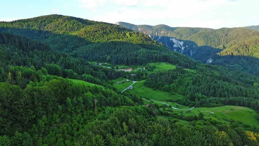 A road passes hills covered with spruce forests and meadows against a cloudy sky