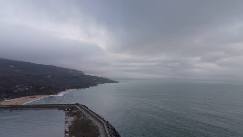 Cinematic aerial view of seaside breakwater under dramatic cloudy sky.