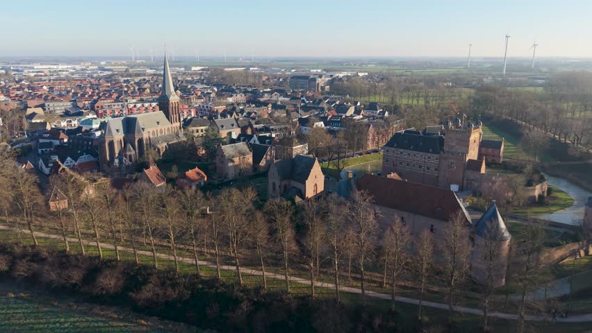 Aerial view of a historic town with a tall church tower, surrounding gabled houses, a castle like complex with a moat, and open fields with wind turbines stretching toward the horizon.