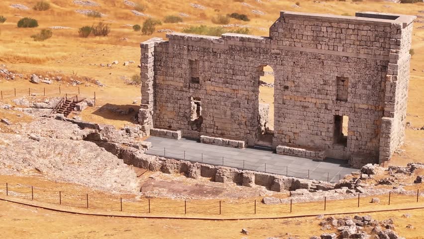Cinematic 4K drone shot of the ancient Roman theater of Acinipo archaeological site, showing the standing stone wall and seating area in the mountains near Ronda, Malaga province.
