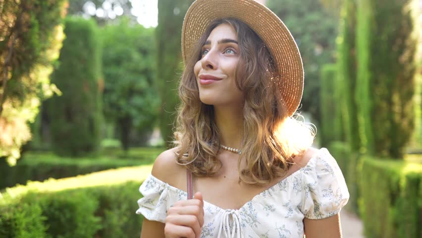 A woman walks along a garden path dressed in a white dress and straw hat. She poses thoughtfully with trees and a building in the background under sunlight.