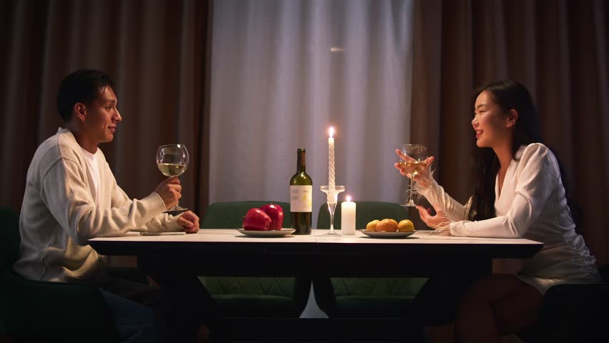Couple have romantic candlelit dinner home, enjoying wine glasses and conversation. Date love night, anniversary celebration, casual evening gathering. Cozy candle light. Man and woman enjoy talking.