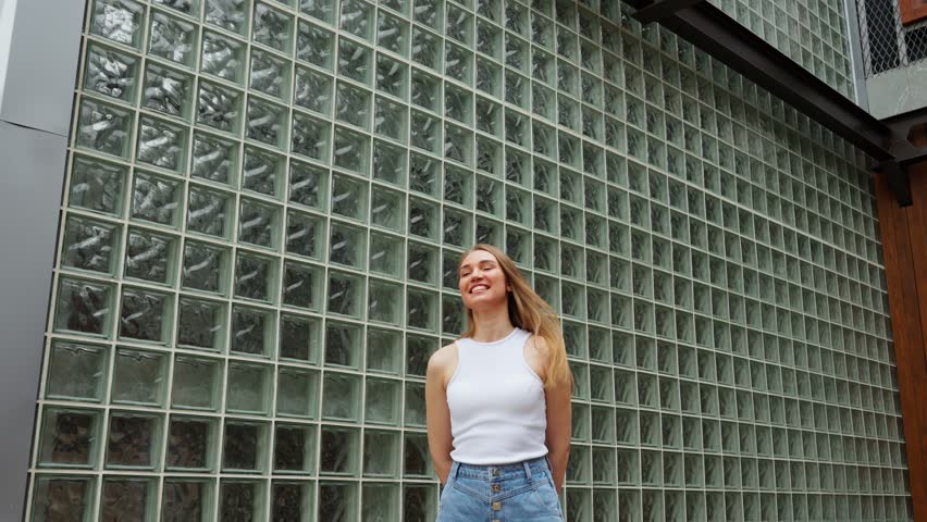 Standing in front of a glass brick wall, a young woman poses casually in a stylish outfit