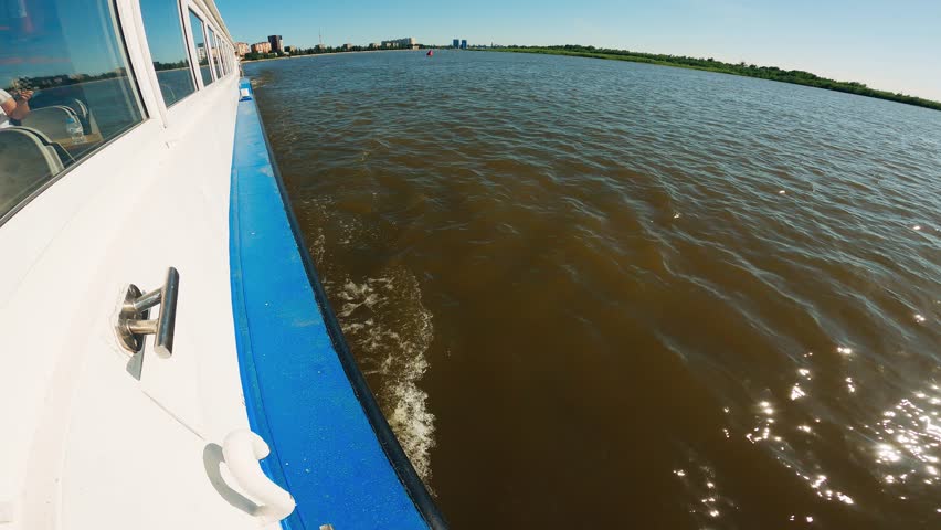 Wide-angle shot from a moving boat on a large river. Bright sunlight sparkles on the murky water. The white and blue vessel creates a wake, with a distant city visible. Serene travel