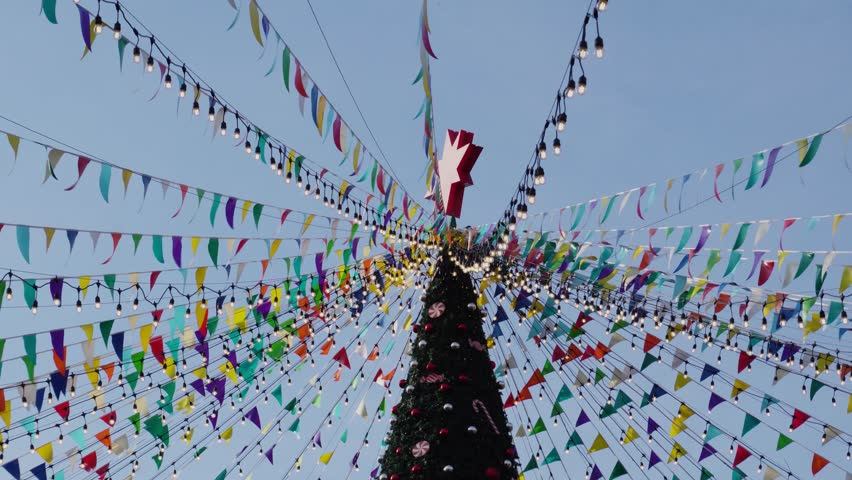 Low Angle View of a Tall Decorated Christmas Tree with Colorful Bunting Flags and Fairy Lights Against Blue Sky