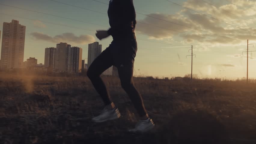 Professional Runner Jogging Across Wild Field At Sunset. Skilled Runner Jogging Through Wild Field During Evening Training Session. Healthy Lifestyle Activity. Runner Jogging In Wild Field Near Urban Skyscrapers Under Golden Sky. Athletic Movement Process. Workout Fitness Exercise. Professional Labor Performance. City Outskirts Journey. Quality Control Speed. Seasonal Daily Workout. Outdoor Recreation Project. Physical Strength Development