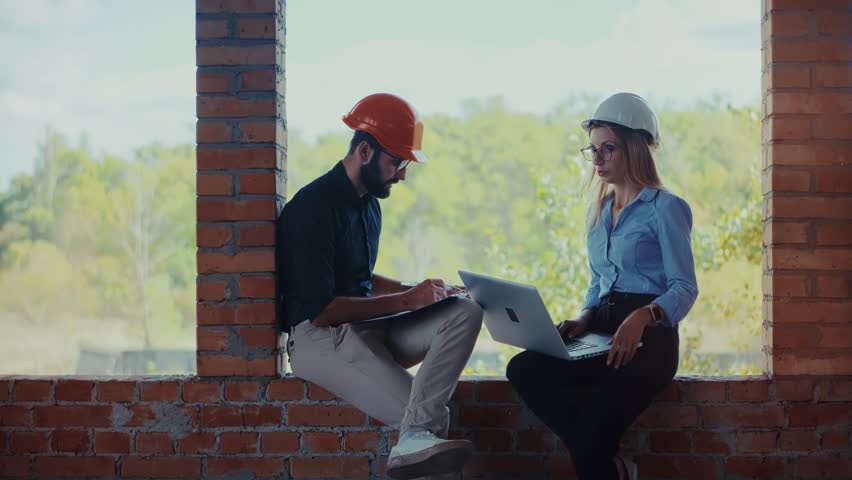 Architects Working With Laptop Computer At Construction Site. Skilled Architects Working Using Laptop Computer To Review Building Project Plans. Industrial Development Activity. Architects Working Together On Laptop Computer While Sitting In Unfinished Brick Structure. Technical Design Process. Engineering Team Labor. House Renovation Service. Property Maintenance Task. Professional Workspace Performance. Urban Improvement Project. Architectural Quality Control. Safety Helmet Equipment