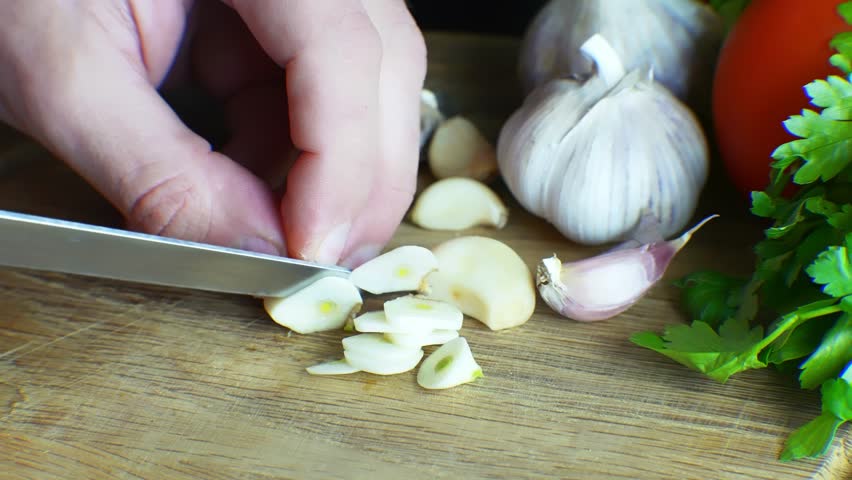 Cook Slicing Fresh Garlic Cloves On Wooden Board. Skilled Cook Slicing Organic Garlic Cloves Using Sharp Knife For Recipe Preparation. Culinary Art Process. Cook Slicing Aromatic Garlic Cloves Near Green Parsley And Red Tomato In Kitchen. Healthy Food Ingredients. Natural Nutrition Cooking. Restaurant Meal Service. Vegetable Cutting Task. Professional Labor Performance. Home Development Menu. Quality Control Inspection. Diet Wellness Style