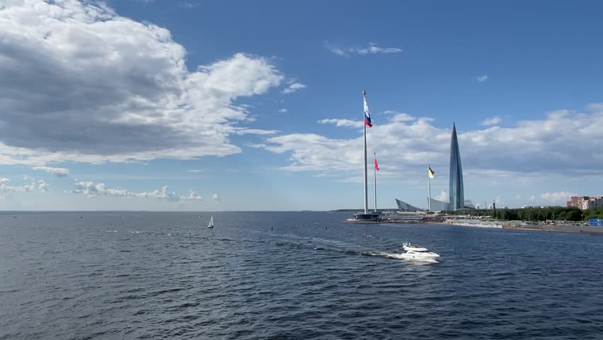 A scenic view of the Gulf of Finland with the iconic Lakhta Center skyscraper dominating the skyline in St. Petersburg, Russia. In the foreground, three national or city flags flutter gracefully in the wind on tall flagpoles along the waterfront. A luxurious yacht cruises calmly across the sparkling blue waters of the Neva Bay, adding a touch of elegance and movement to the tranquil summer seascape. The clear sky and gentle clouds complete this picturesque urban coastal scene, showcasing the blend of modern architecture, maritime activity, and natural beauty. Footage perfect for travel, city life, luxury lifestyle, and Russian tourism themes.