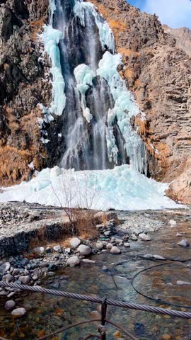 Experience the serene beauty of a frozen waterfall in a rugged mountain landscape. This breathtaking scene captures the icy blue hues of a partially frozen waterfall, set against a backdrop of rocky terrain and a clear blue sky. Ideal for use in projects evoking a sense of natural wonder, adventure, and tranquility.