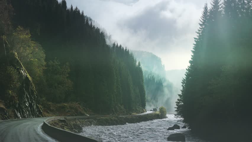 A wild mountain river rushes along the narrow road. The steep forest-covered mountains on both sides. Thin fog in the air.