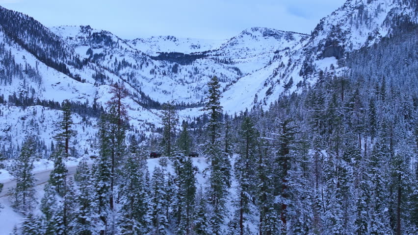 Drone aerial of vehicles driving along Emerald Bay Road through snowy alpine forest as soft snow falls. Curving mountain highway above Lake Tahoe in winter conditions. Emerald Bay, Lake Tahoe, CA, USA