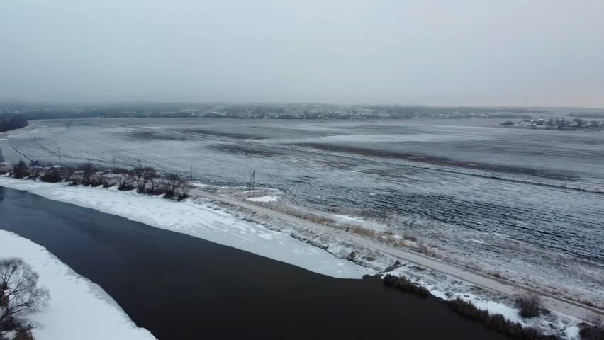 Aerial view. Cloudy weather covers a rural area where a river flows calmly beside icy fields. The landscape shows trees and patches of snow along the riverbank.