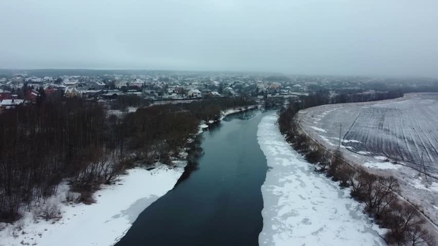 Aerial view. A view of a river flowing through a town covered in snow. Ice is present along the riverbanks. The scene shows the winter weather with cloud cover and bare trees.