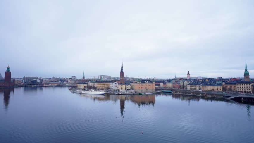 Stockholm skyline across Riddarfjärden with Riddarholmen Church and Stockholm City Hall reflected on calm water under an overcast sky, Sweden.