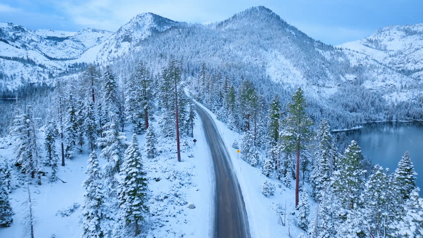 Aerial view of Emerald Bay Road cutting through snow covered pine forest with mountain peaks in background. Winter alpine landscape near Lake Tahoe, California, USA.