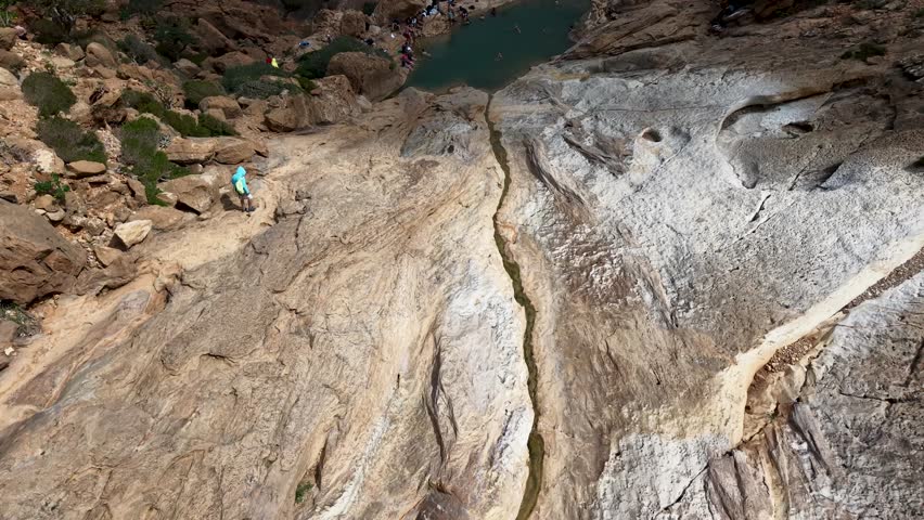 Aerial view of hikers and swimmers at a natural rocky pool in a canyon, with green water surrounded by rugged stone terrain, outdoor adventure and summer recreation.
