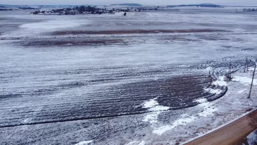 A view shows a snowy field with tire tracks on the ground. Distant hills are visible. The scene captures the winter landscape in a rural area. The sky appears cloudy.