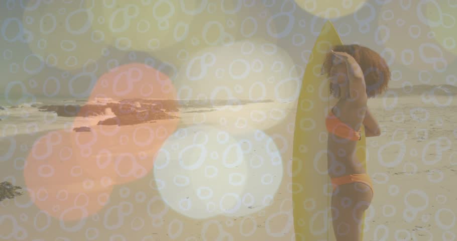 Woman with yellow board scanning waves, adjusting hair prepping to surf, bokeh overlay warming her. Beach, surfer, sand, swimwear, sunlit, horizon, serene