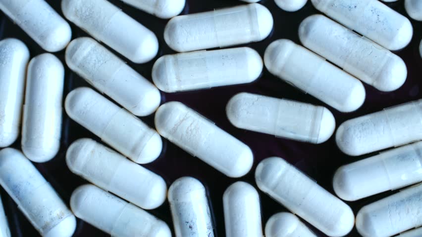 Close up numerous white medical capsules containing powder, rotating on dark background, illustrating concepts of pharmacology, addiction, and healthcare. White pharmaceutical supplement capsules.