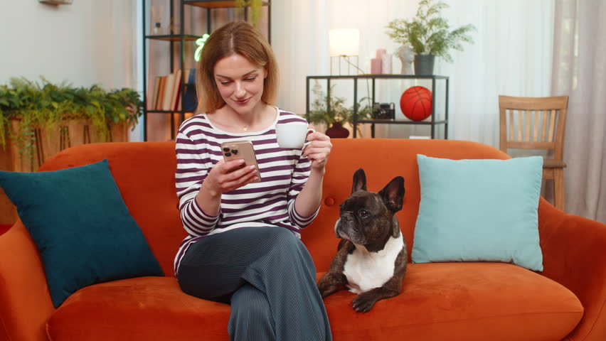 Middle-aged woman drinks coffee with French bulldog at home reads news on smartphone feels calm and happy. Girl pets dog on sofa smiles enjoys mug morning browsing news app lifestyle daily online feed
